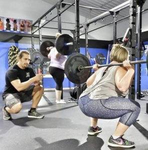 A woman performs a squat at the bar under the supervision of an Elite Edge coach.